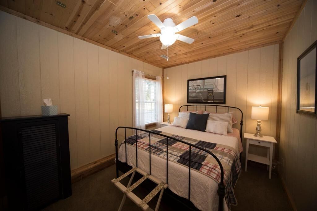 A bedroom with a bed, a ceiling fan, a nightstand, a lamp, and a window with curtains.
