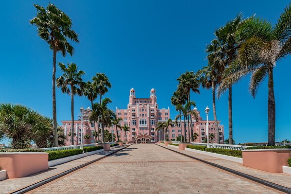 A large, pink, multi-story hotel with a central entrance, surrounded by palm trees and a paved walkway.