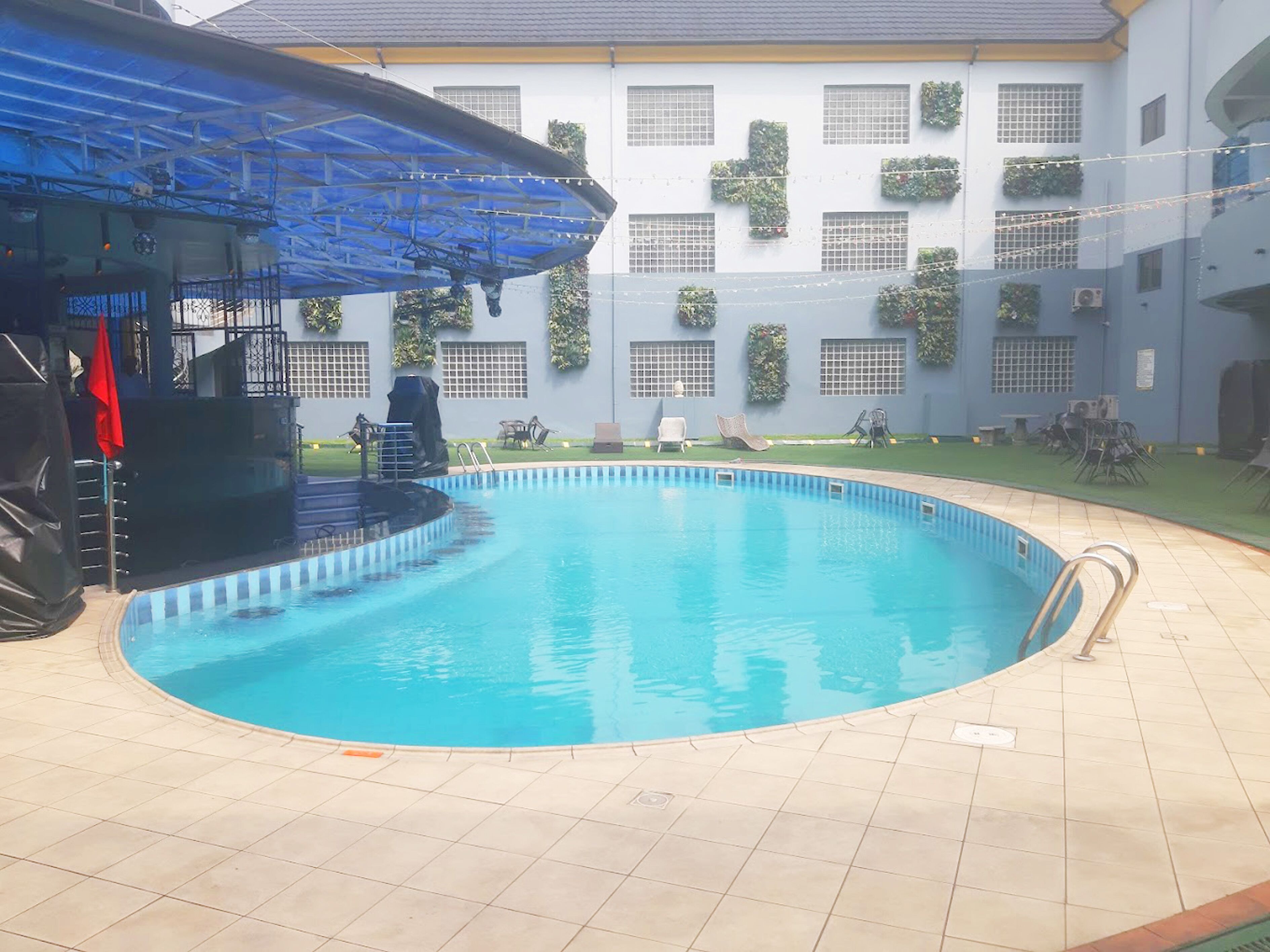 A swimming pool with a blue-tiled deck, surrounded by a covered area with chairs and a barbecue grill.