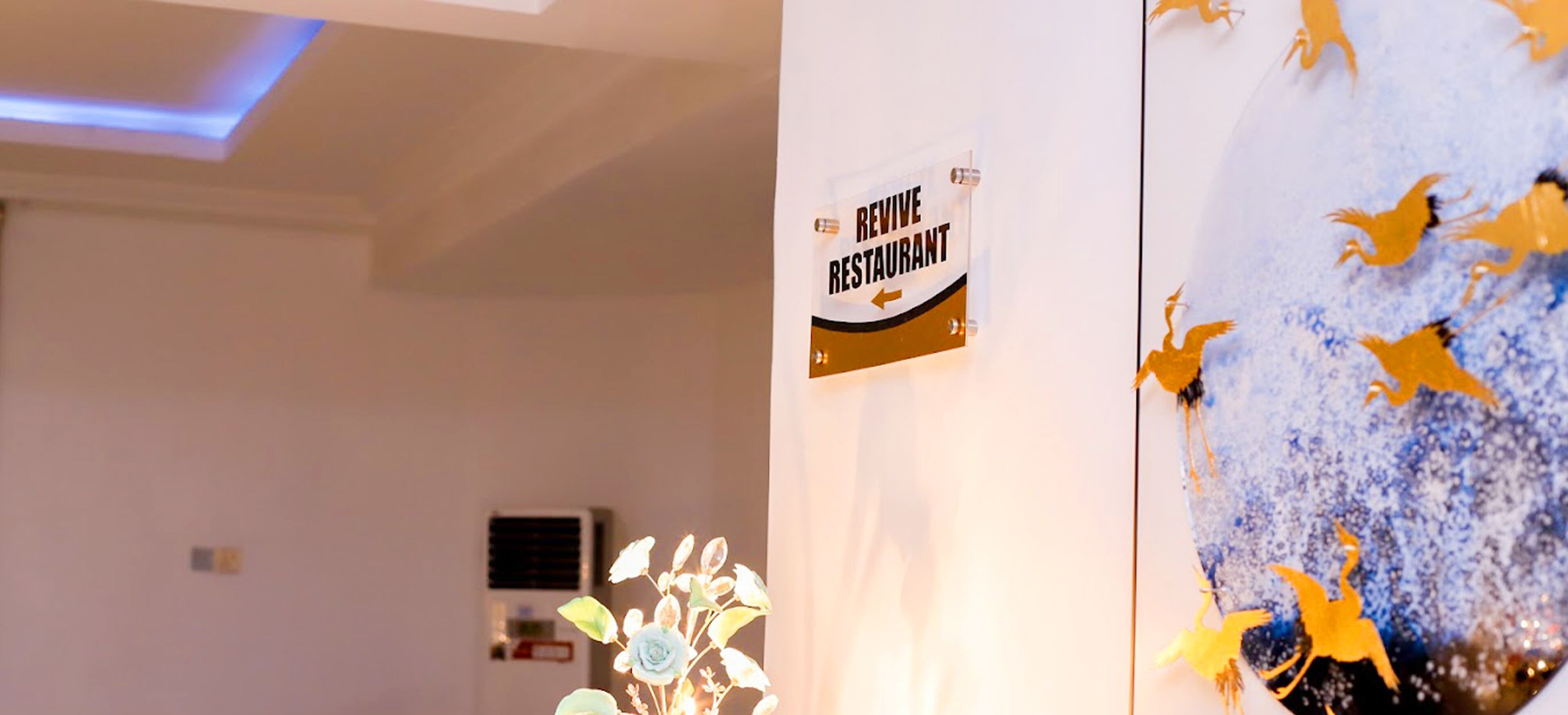 A sign for 'Revive Restaurant' with a directional arrow, a decorative wall with golden figures, and a ceiling with blue lighting.