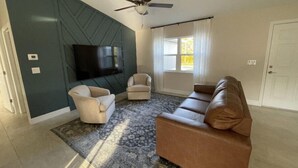 A living room with a ceiling fan, a flat-screen TV mounted on a dark wall, two armchairs, a brown leather sofa, and a patterned rug. - Charming, Modern, Cozy Retreat (North Port)