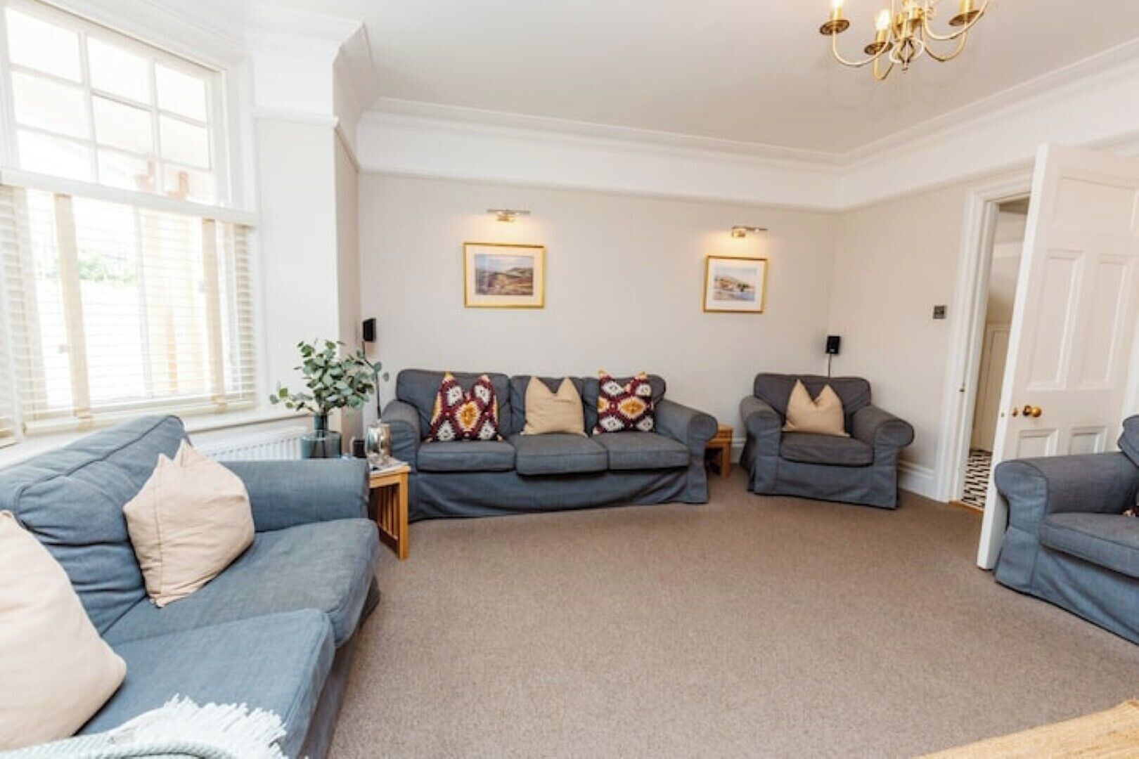 A living room with two grey sofas, a coffee table, and a chandelier. There are two framed pictures on the wall and a door to the right.