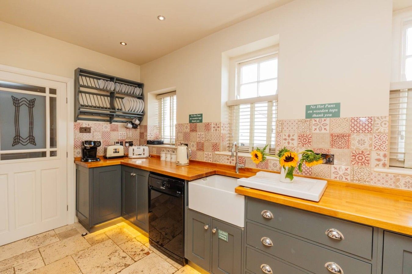 A kitchen with a wooden countertop, a dishwasher, and a sink. There are two windows with blinds, and a sign on the wall.