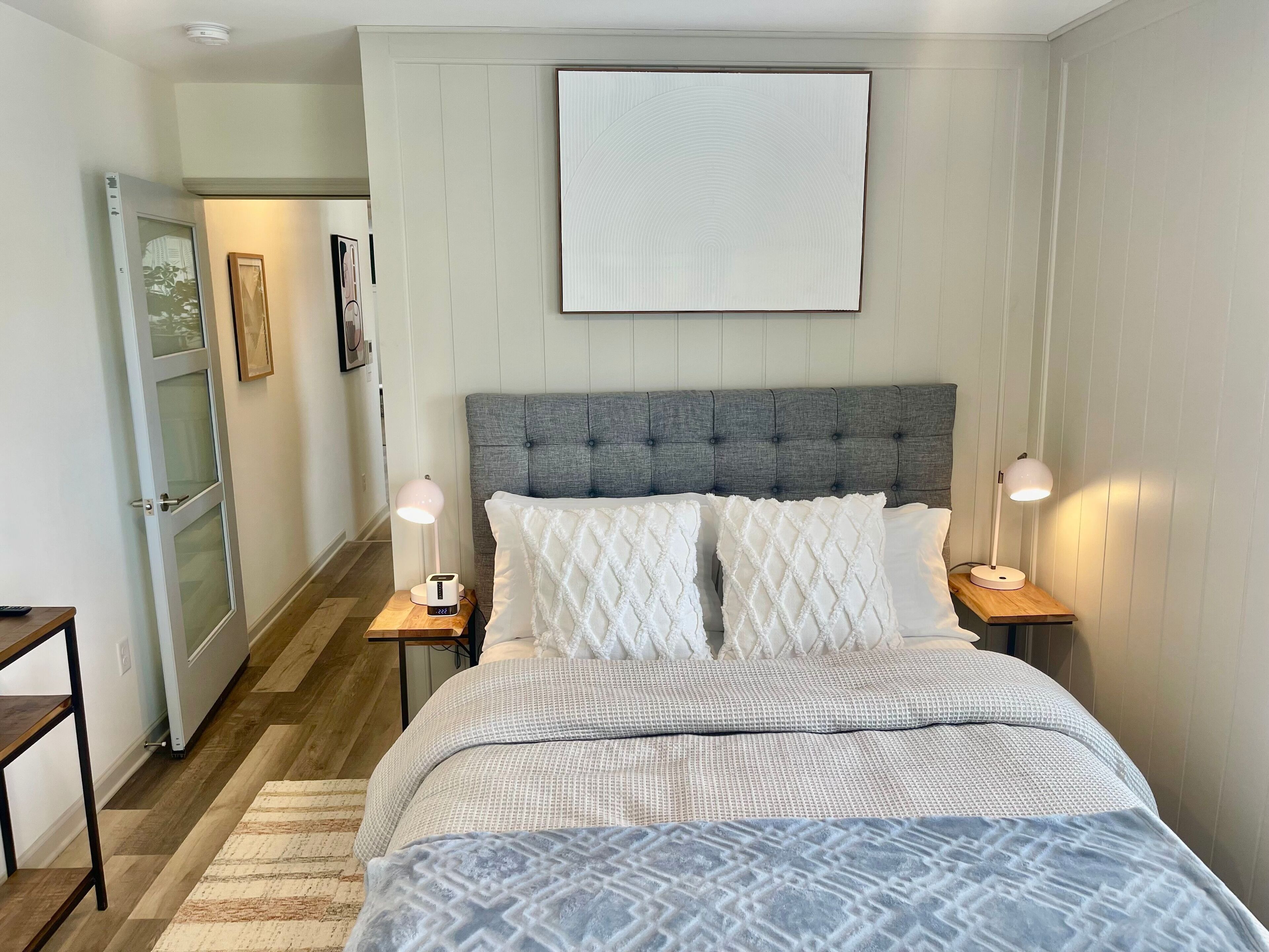 A bedroom with a bed, two bedside tables, a nightstand, a door, and a wall-mounted artwork.