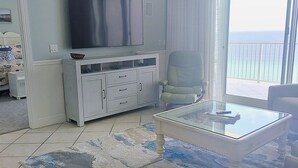 A living room with a flat-screen TV, a glass coffee table, a sofa, and a view of the ocean through a large window. - 1204 - Free Beach Chairs and Amenity Package! (Panama City Beach)