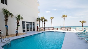 A swimming pool with a view of the beach, surrounded by palm trees and lounge chairs. - 1204 - Free Beach Chairs and Amenity Package! (Panama City Beach)