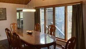 A dining room with a large round table, wooden chairs, a chandelier, and large windows with curtains.