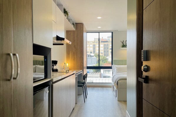 A compact hotel room with a kitchenette, a bed, a window with a city view, and a door with a lock.