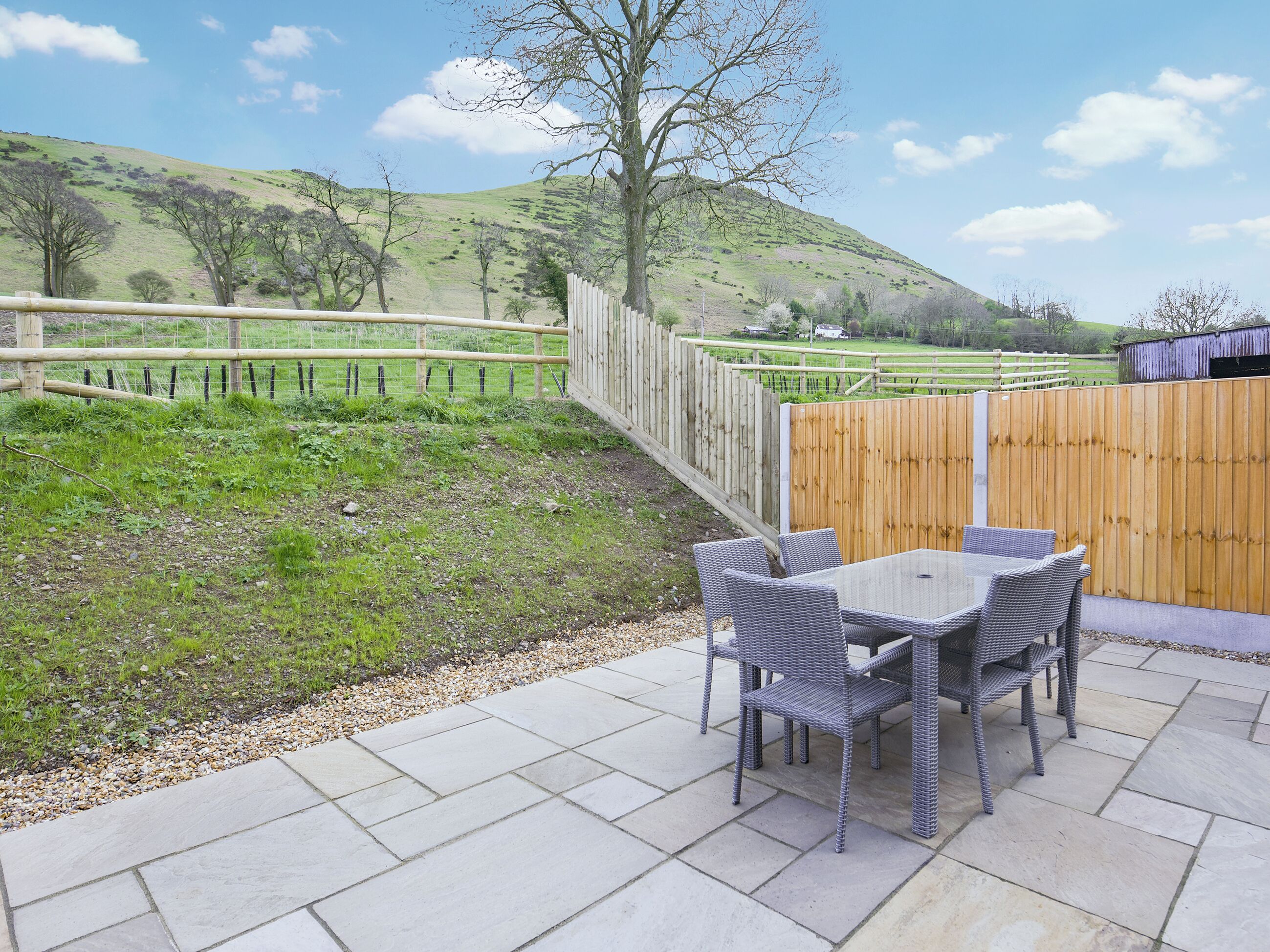 An outdoor patio with a table and chairs, a wooden fence, and a view of a hilly landscape.