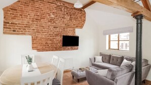 A room with a brick wall, a television mounted on it, a grey sofa, a white dining table with chairs, and a window with a view of buildings. - 3 bedroom accommodation in Church Stretton (Church Stretton)