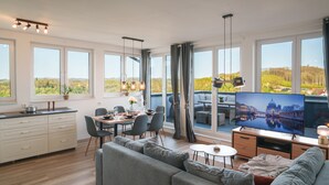 Un salon moderne doté d’un grand téléviseur à écran plat, d’un canapé gris, d’un coin repas avec des chaises et d’une cuisine avec des meubles blancs.