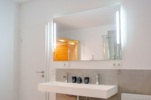Ein modernes Badezimmer mit einem weißen Waschbecken, zwei Wasserhähnen und einem großen Spiegel.