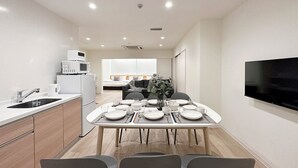 A modern kitchen with white cabinets, a built-in microwave, and a sink. There is a dining area with a table set for four, chairs, and a flat-screen TV mounted on the wall. - Nikke 201 (Hiroshima)