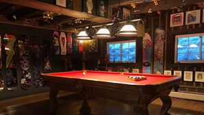 Une table de billard dans une pièce dont le sol et les murs sont en bois, et qui est décorée de planches à roulettes et de tableaux encadrés.