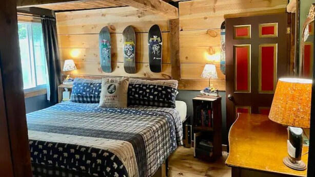 A cozy bedroom with a wooden ceiling, a bed with patterned bedding, two wall-mounted skateboards, a nightstand with a lamp, and a door with red panels.