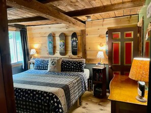 A cozy bedroom with a wooden ceiling, a bed with patterned bedding, two wall-mounted skateboards, a nightstand with a lamp, and a door with red panels. - Smugglers' Notch - Grunberg Haus Inn & Cabins (Waterbury)
