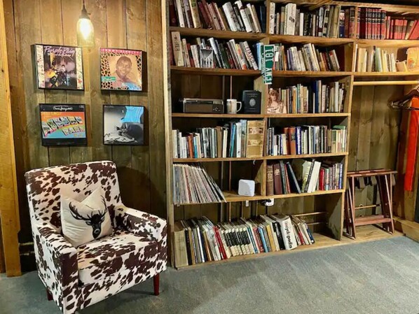 Una habitación con una estantería de madera llena de libros y CDs, un sillón estampado con vacas y portadas de discos enmarcadas en la pared.