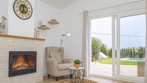 Ein Wohnzimmer mit Kamin, einem gemütlichen Sessel, einem kleinen Tisch mit Pflanze und Blick auf einen Pool durch große Schiebetüren.