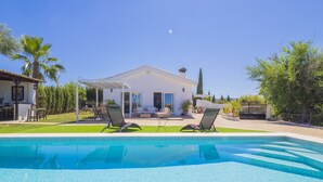 Ein Schwimmbecken mit Liegestühlen, eine Terrasse und der Blick auf ein Haus mit weißer Fassade und einer überdachten Terrasse.
