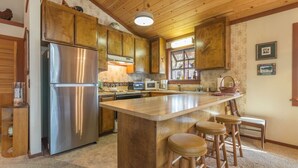 A kitchen with wooden cabinets, a stainless steel refrigerator, a central island with stools, and a window with decorative items. - Beautiful Cabin In Tahoe Vista! (Tahoe Vista)