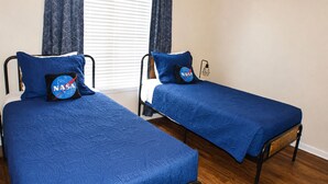 Two single beds with NASA-themed pillows, a window with blinds, and a wall-mounted lamp. - Space Oddity: Close & Accessible! / Fiber Internet / EV Charger! (Huntsville)