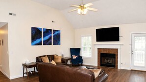 A living room with a brown leather sofa, a blue armchair, a flat-screen TV, a fireplace, and three wall-mounted paintings. - Space Oddity: Close & Accessible! / Fiber Internet / EV Charger! (Huntsville)