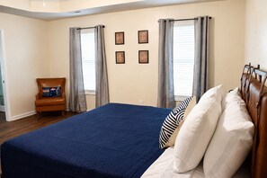 A bedroom with a large bed, two windows with curtains, a chair with a pillow, and wall art.