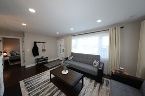 Una sala de estar moderna con un sofá gris, una mesa de centro con objetos decorativos, un banco negro y una mesita de noche de madera.