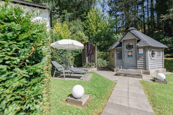 A wooden cabin with a small porch, a covered seating area with chairs and an umbrella, and a stone pathway leading to the cabin.