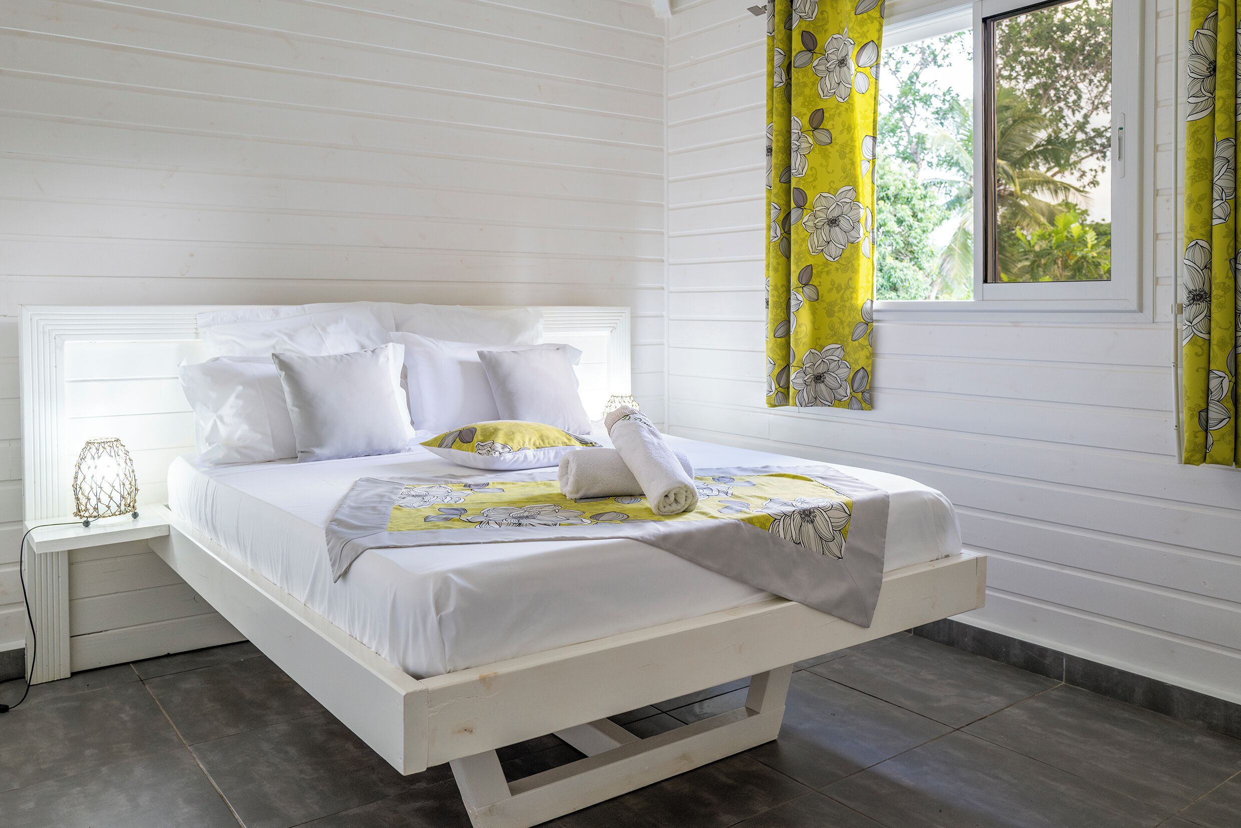 A white wooden bed with white bedding, a bedside table with a lamp, and a window with curtains.
