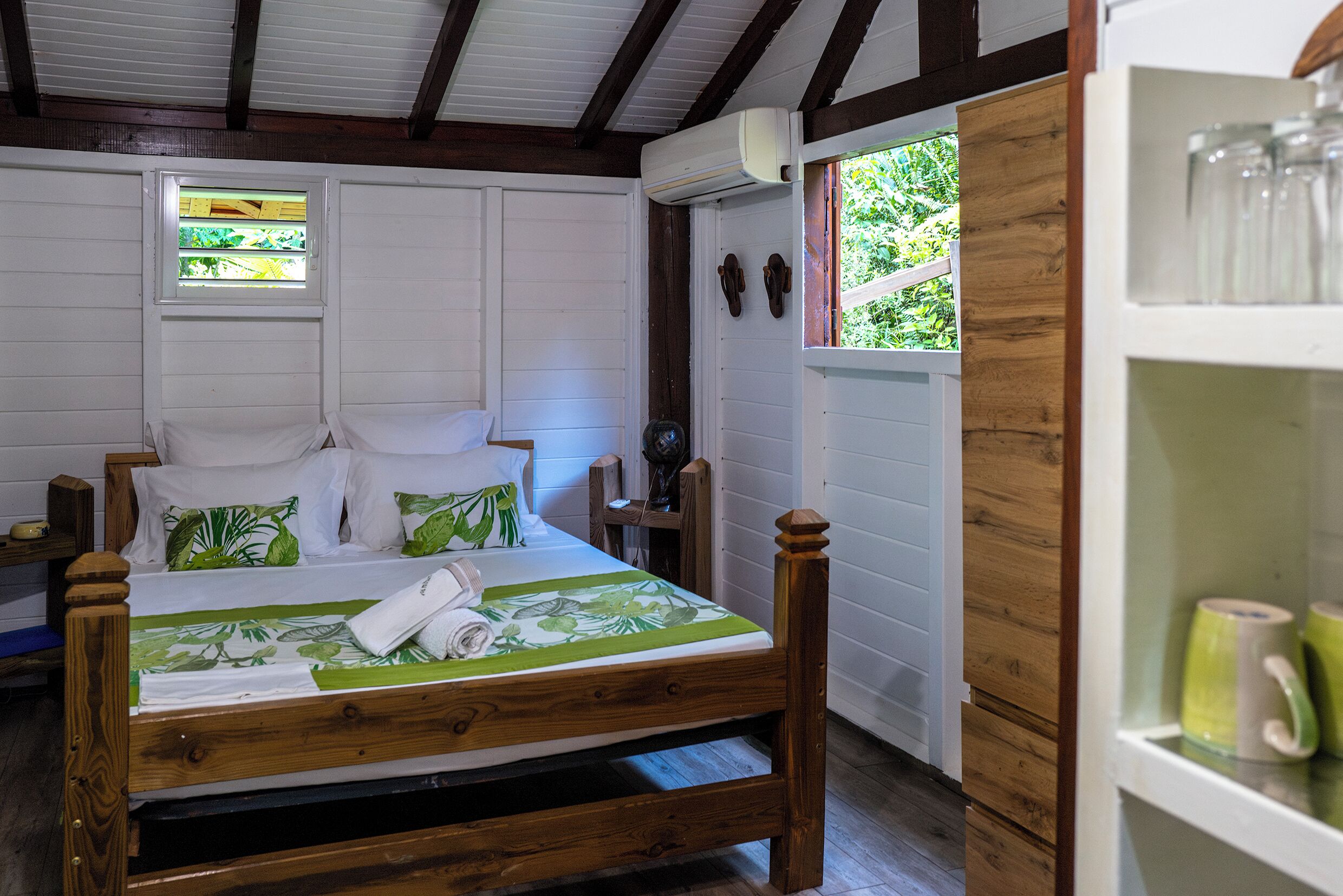 Een houten bed met een beddegoed met een groen bladpatroon, een wit kussen en een houten hoofdbord. Er is een klein raam met een geel luik en een witte airconditioner boven het bed.