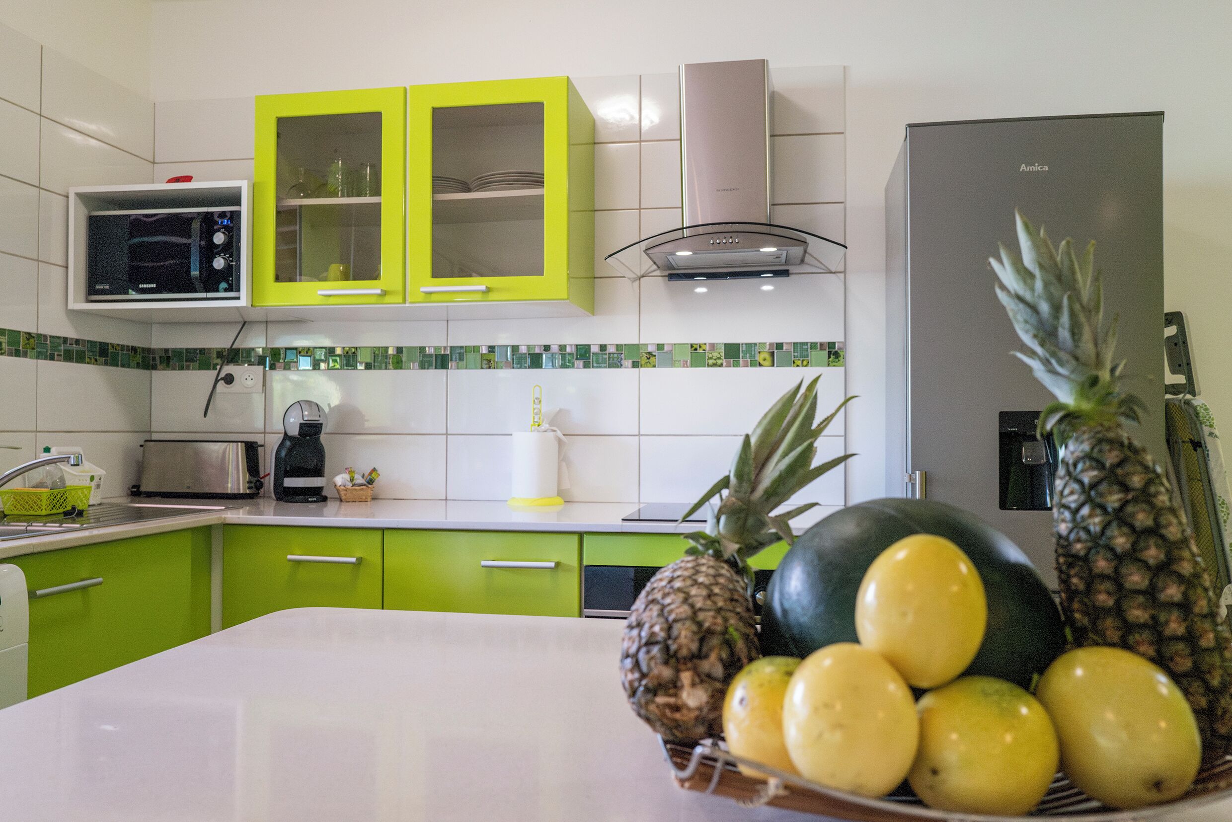 Une cuisine avec des meubles de rangement verts, un four à micro-ondes, un grille-pain et un panier de fruits contenant de l’ananas, du melon et des citrons.