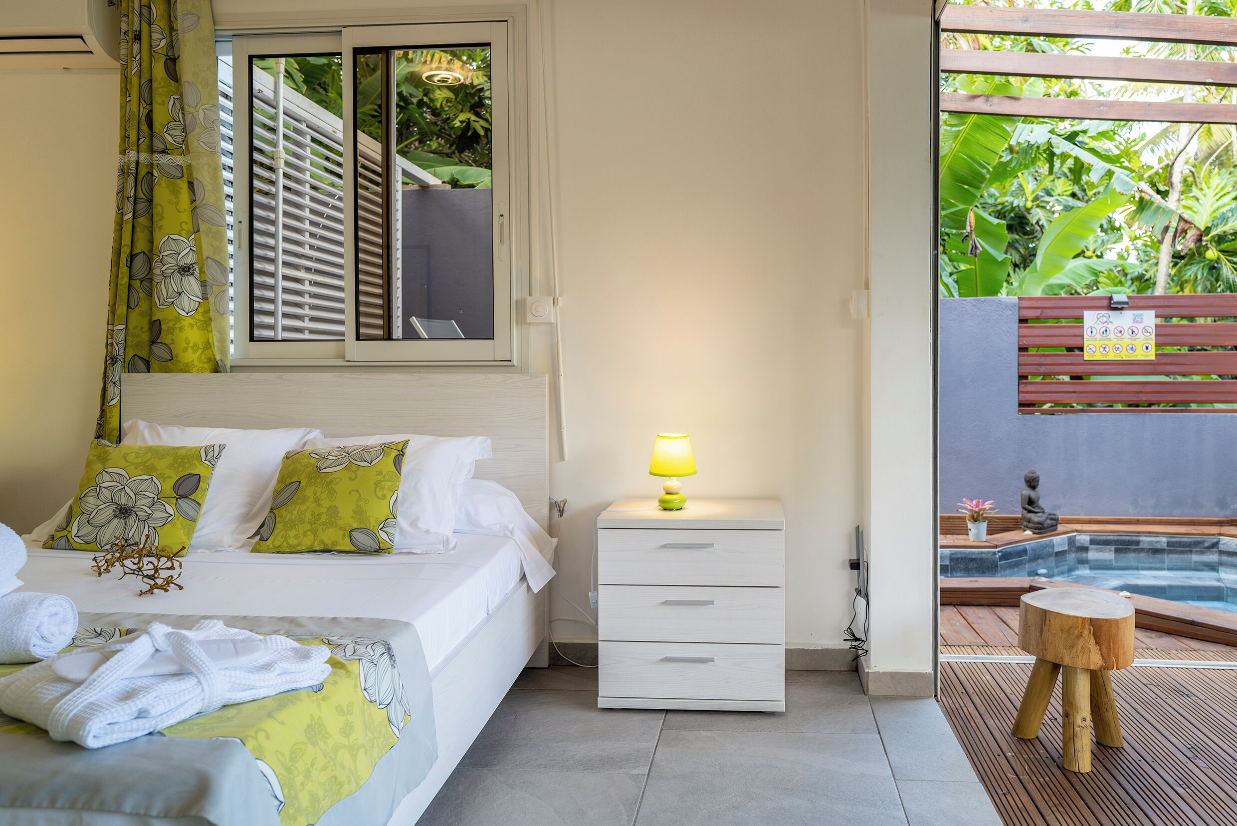 Une chambre d’hôtel avec un lit, une table de chevet avec une lampe, un tabouret en bois et une vue sur une piscine extérieure.