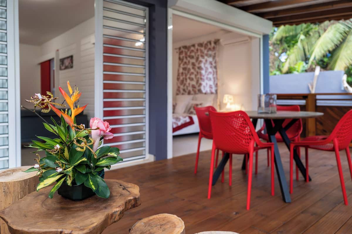 A wooden dining table with red chairs, a potted plant with flowers, and a room with a bed and a window with blinds.