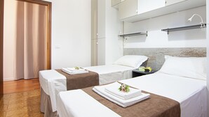 A hotel room with two beds, each with a white bedspread and a brown headboard. There is a small table with a lamp and a book on it.