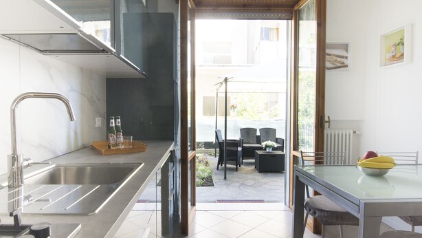 A modern kitchen with a stainless steel sink, a dining area with a glass table and chairs, and a view of the outdoor patio.