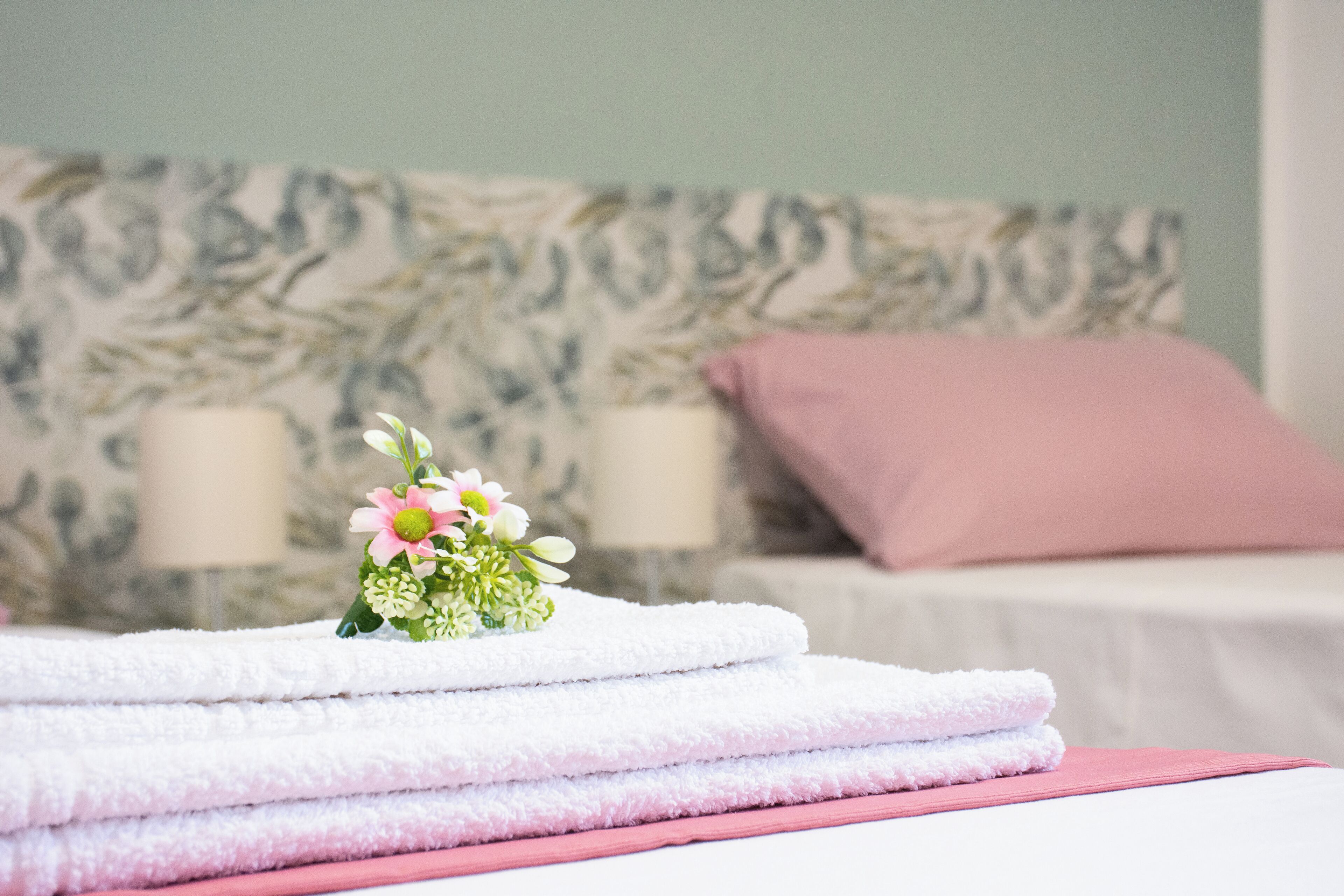 A neatly folded stack of white towels with a small floral arrangement on top, placed on a bed with a pink pillow and a floral-patterned headboard.