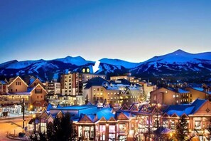 A town with a mix of traditional and modern buildings, a prominent clock tower, and snow-capped mountains in the background.