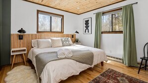 Ein Schlafzimmer mit Holzbalkendecke, einem großen Bett mit weißer Bettwäsche, zwei Nachttischlampen, einem Fenster mit grünen Vorhängen und einem Teppich auf dem Boden.