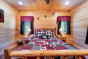 Una habitación de cabaña acogedora con interior de madera, una cama con manta de cuadros, dos mesitas de noche con lámparas y un ventanal con cortinas.
