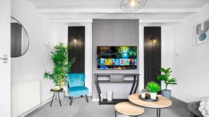 A modern living room with a flat-screen TV mounted on the wall, a blue armchair, a small round table with a plant, and a grey sofa.