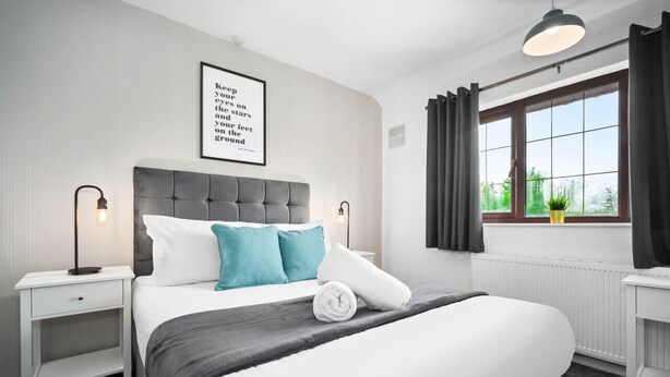 A neatly arranged bedroom with a large bed, a bedside table, a nightstand, a window with curtains, and a wall-mounted light fixture.