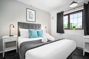 Un dormitorio ordenado con una cama grande, una mesita de noche, cortinas en una ventana y una lámpara fijada a la pared.