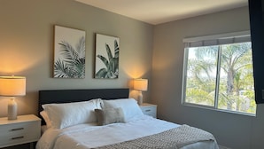 A bedroom with a bed, two nightstands, two wall-mounted lamps, a window with a view of palm trees, and two framed botanical prints on the wall. - 5-10 mins to Temecula WineCountry; resort-like pool spa w/ amazing sunset views! (Winchester)