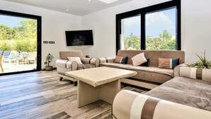 Ein modernes Wohnzimmer mit einer Couch, Sesseln und einem Couchtisch. Ein Fernseher ist an der Wand befestigt und große Fenster bieten einen Blick ins Freie.