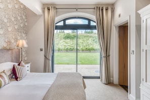 Quarto com cama, mesa de cabeceira, abajur e vista para um jardim através de uma porta de vidro.