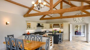 A spacious kitchen with wooden beams, a dining area with a wooden table and chairs, and a view into a living area with a sofa and a clock on the wall.