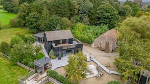 A modern house with a large deck, surrounded by lush greenery and a stone barn.