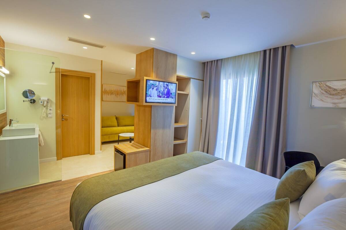A hotel room with a bed, a TV mounted on a wooden panel, a small desk, a sofa, a bathroom with a sink and mirror, and a shower area.