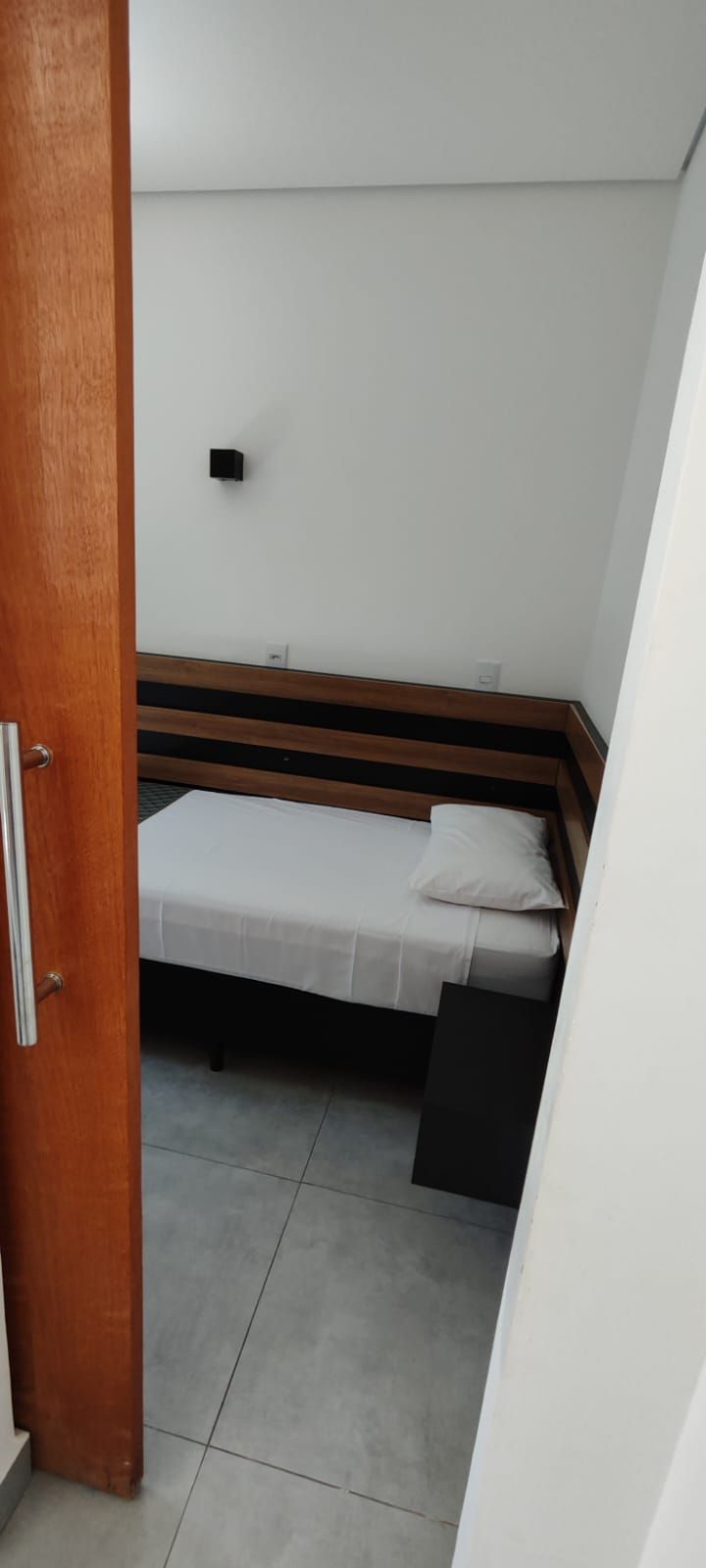 Una habitación pequeña y estrecha con una cama individual, cabecera de madera y pared blanca.